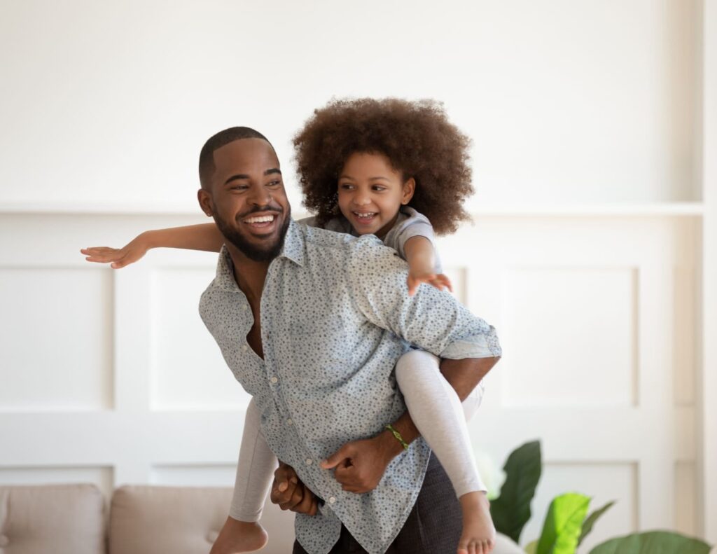 Smiling dad gives daughter a piggyback ride at home, highlighting joyful moments made possible by hearing wellness.